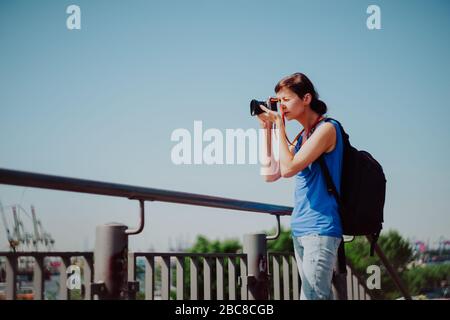 Attractive young female tourist with backpack and camera is exploring Hamburg city. Woman is making photo on retro camera and looking for new motives. Stock Photo