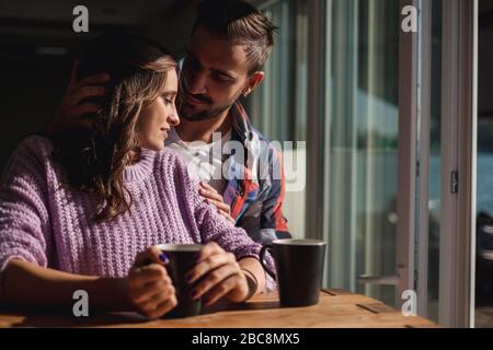 Handsome man hugging girlfriend while they drink coffee by the window on a beautiful sunny day Stock Photo