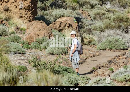 A young man in a cap with a backpack behind his back travels along a route in the Teide National Park. Hiking by the mountain trail surrounded by ende Stock Photo
