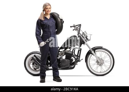 Full length portrait of a female mechanic standing next to a custom motorbike and holding a tire and a wrench isolated on white background Stock Photo