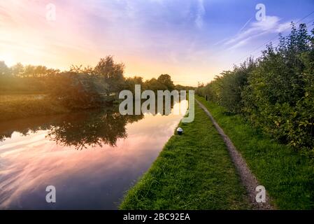 Looking along an English canal tow path on a summer evening as the sun sets with a beautiful sky reflecting in the water Stock Photo
