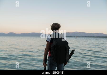 View from behind of a young male photographer standing by the evening sea with photographic backpack on his back and tripod in his hands. Stock Photo