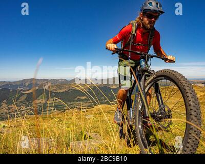 Mountain bikers on the single track GR 400 below the summit of the Plomb du Cantal. View of the Vallée de la Cère valley. In the background the Puy Gr Stock Photo