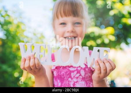 Little toddler - laughing girl in a pink dress holding the words WELCOME in her hands Stock Photo