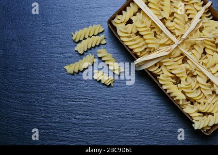 Background and space for text with a cardboard box of raw spiral noodles photographed from above, on a gray slate board and a bow made of bast Stock Photo