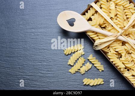 Background and space for text with a cardboard box of raw spiral pasta photographed from above, on a gray slate board and a bow made of bast and a woo Stock Photo