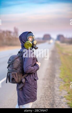 A girl in a black gas mask stands on the edge of a suburban highway. The girl is trying to stop passing cars in order to leave the city in which the e Stock Photo