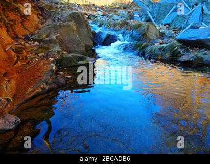 A stream with a red dirt bank on one side and large blue stones with green moss on the other side bubbles through the rocky terrain. Stock Photo