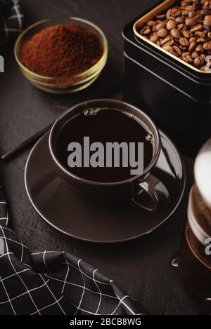 A black coffee Cup,a checkered towel, a metal jar of roasted coffee beans on a dark concrete table Stock Photo