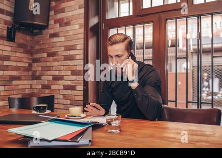 Successful bearded business man writing document during mobile phone conversation with client, sitting at table in cafe. Male skilled financier having