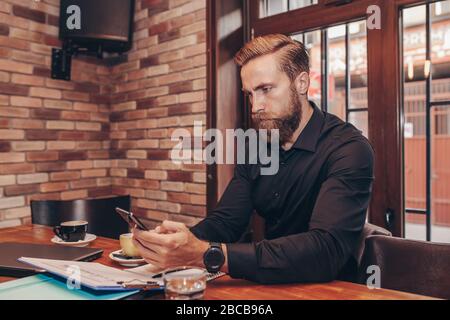 Handsome bearded businessman working with smartphone and documents, while sitting in cafe. Stock Photo