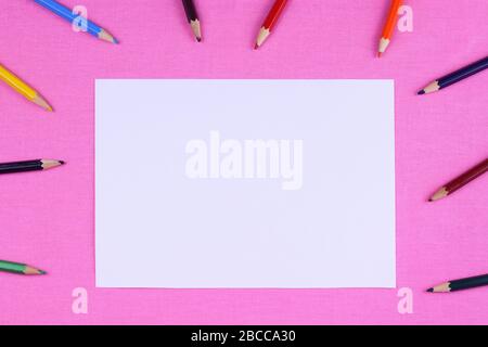 Multicolored pencils for drawing around empty white paper on a pink table. Place for text Stock Photo