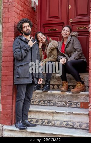 Istanbul, Turkey - February 12, 2020: Smiling young people from Balat quarter of Fatih district are sitting on the steps at the red front door of the Stock Photo