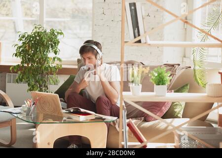 Young man studying at home during online courses for marketer, architect, translator. Getting profession while isolated, quarantine against coronavirus spreading. Using laptop, smartphone, headphones. Stock Photo