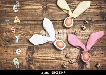 Beautiful Easter background with eggs in napkins like Easter Bunny and quail eggs on rustic wooden table. Top view with space for text. Stock Photo