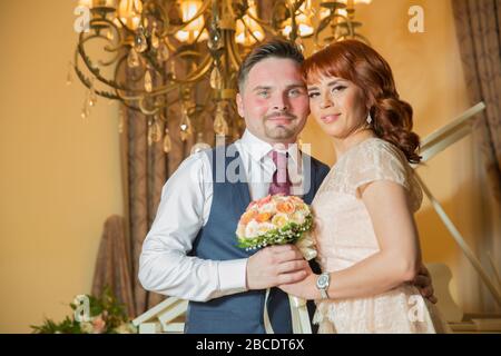 Bridal couple in front of a piano . Happy wedding day. Happy bride and groom near piano in white interior studio . Newlyweds stand in the interior Stock Photo