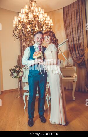 Bridal couple in front of a piano . Happy wedding day. Happy bride and groom near piano in white interior studio . Newlyweds stand in the interior Stock Photo