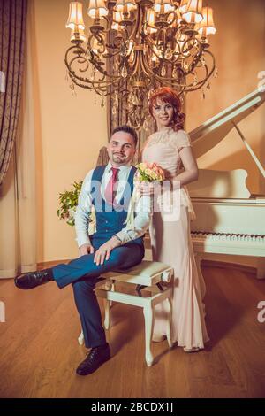 Bridal couple in front of a piano . Happy wedding day. Happy bride and groom near piano in white interior studio . Newlyweds stand in the interior Stock Photo