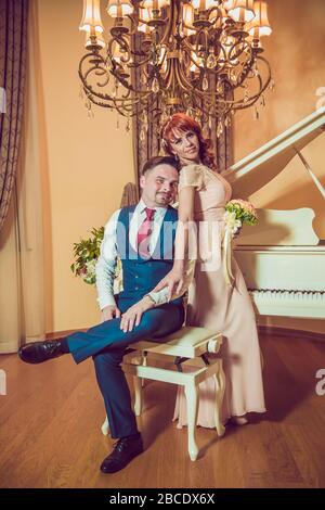 Bridal couple in front of a piano . Happy wedding day. Happy bride and groom near piano in white interior studio . Newlyweds stand in the interior Stock Photo