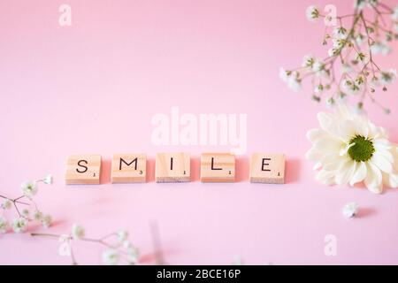 The word SMILE in wooden scrabble style tiles laid out on a pink background with white flowers - still life concept image with copy space. Stock Photo