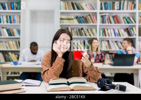 Cheerful pretty smart girl student in casual wear, looking at camera and smiling, holding cup of coffee or tea and preparing for exams in university Stock Photo