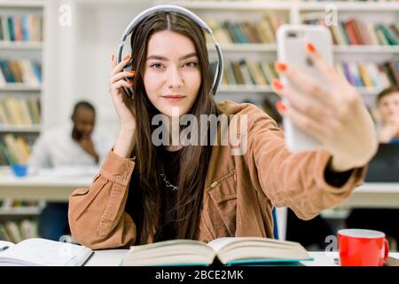 Pretty Caucasian young woman student in casual hipster clothes, making selfie photo on smartphone for sharing it in networks, sitting at the table Stock Photo