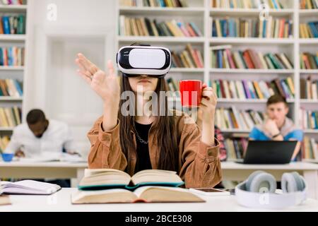Young smart confident woman in casual clothes sitting in library and experiencing new device VR headset for study, touching the air and holding red Stock Photo
