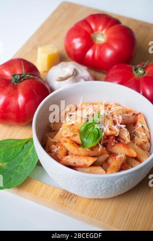 Classic italian penne pasta arabiata with its ingredients in the background: fresh tomatoes, basil, garlic and parmesan cheese Stock Photo