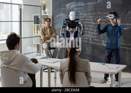 Teenage student with vr headset standing by blackboard in front of his classmates and demonstrating abilities of robot near by Stock Photo
