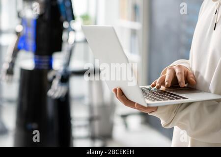 Hand of teenager with laptop developing new software while working over technological project on background of robot in college Stock Photo