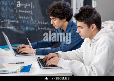 Two teenage guys typing in front of laptops while sitting by desk at lesson during individual work on background of blackboard Stock Photo