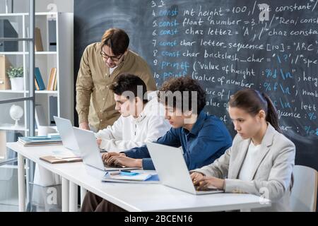 Young teacher checking individual work of one student and consulting him while other classmates preparing their presentations near by Stock Photo