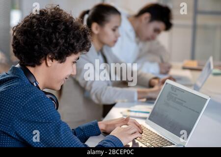 Successful teenage student looking at laptop display while entering information during work over technical project at lesson Stock Photo