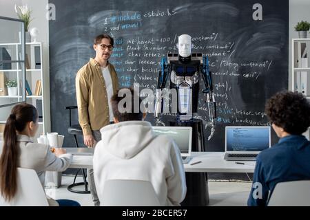 Young teacher standing by blackboard in front of audience and making presentation of robot to group of students at lesson Stock Photo