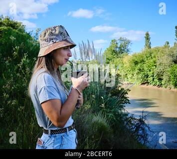 a beautiful girl model drinking yerba mate hot infusion beside the river in Bel Ville City, Cordoba, Argentina Stock Photo