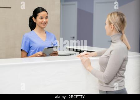 Pretty young consultant with tablet standing by reception counter in front of female patient and registrating her for medical appointment Stock Photo