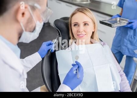 Young smiling blond female patient of dentistry clinics sitting in armchair and looking at her dentist with hook and mirror before oral checkup Stock Photo
