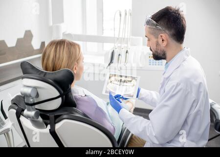 Young male dentist in gloves and whitecoat explaining his patient all the problems with her teeth while showing x-ray in tablet Stock Photo