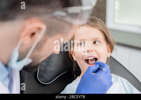 Cute little girl with open mouth looking at dentist in gloves bending over her during oral checkup with mirror in dental clinics Stock Photo