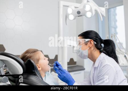 Young female dentist in whitecoat, protective eyeglasses, mask and gloves going to do oral checkup of oral cavity of her little patient Stock Photo