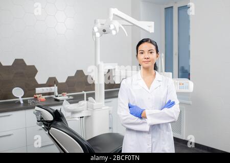 Young confident blunette female dentist in whitecoat and gloves crossing arms by chest while standing in front of camera in dental clinics Stock Photo