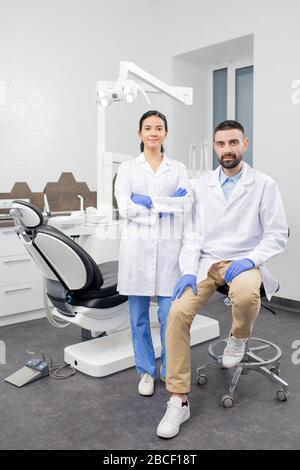 Two young contemporary dentists in gloves and whitecoats looking at you by their workplace in dental clinics with medical equipment behind Stock Photo