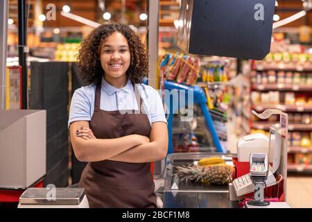 Pretty young smiling African-american female sales clerk with her arms crossed by chest looking at you by cash register during work Stock Photo