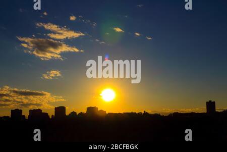 Lovely sunset, with golden sun, with gray-white clouds, with azure sky, with lens flares against the background of the city silhouette. Stock Photo