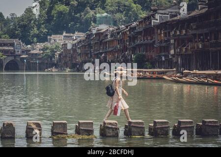 Feng Huang, China -  August 2019 : Chinese lady in wicker hat crossing waters on stepping stones on Tuojiang river, flowing through the centre of Feng Stock Photo
