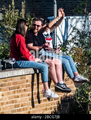 London, UK. 4th Apr 2020. A sunny day and flatmates with drinks climb onto the wall of the roof of a neighbours house to get the last rays of sunshine. The 'lockdown' continues for the Coronavirus (Covid 19) outbreak in London. Credit: Guy Bell/Alamy Live News Stock Photo