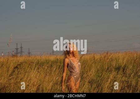 Young woman in golden glittering bodysuit among the field on sunset, power lines in background