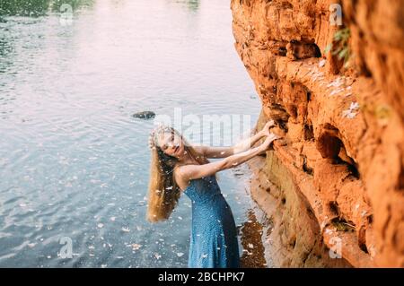 Young woman with closed eyes in crown from shells and threw back head staying near red sand wall in river Stock Photo