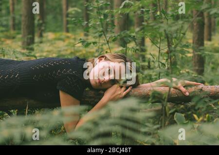 Portrait of young beautiful girl in black dress in forest that lies on the tree trunk, looking to the camera Stock Photo