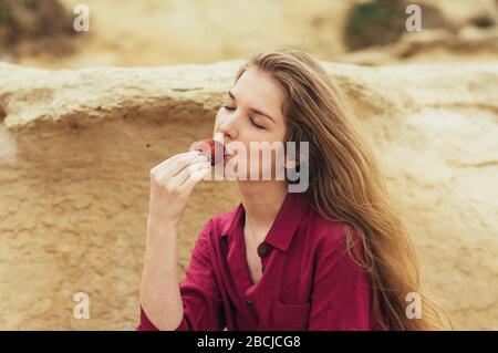 Beautiful blond girl in purple dress eating fresh strawberry on nature Stock Photo